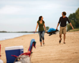 Q 1200 Gas Grill - Blue with a man and woman carrying it on a beach.