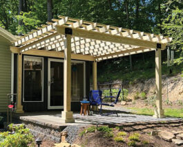 Country Lane Hollander wooden pergola on concrete patio up against green vinyl house with landscaping surrounding.